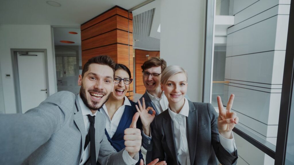 Four colleagues in suits cheerfully posing for a group selfie indoors.
